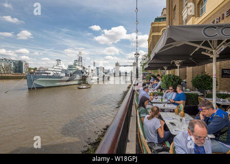 A busy restaurant terrace overlooking the river thames and HMS Belfast at the tourist hotspot area on the South Bank in Central London in summer Stock Photo