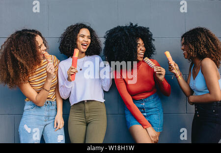 Friends leaning to a grey wall enjoying eating ice cream outdoors. Group of cheerful buddies eating ice-cream. Stock Photo
