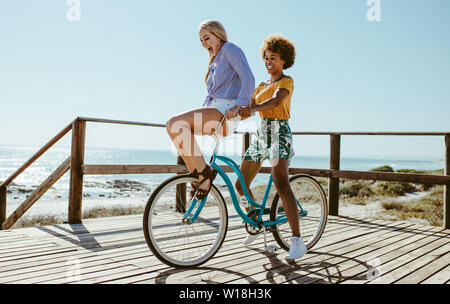 Young woman balancing on the handlebars of a bicycle ridden by her friend on a summer day at the beach. Friends going on a bike ride at the beach. Stock Photo