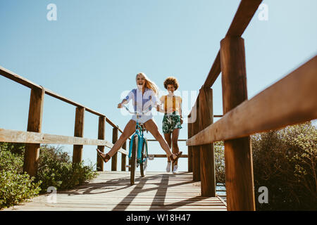 Excited woman riding bike down the boardwalk with her friends running behind. Two young female friends having a great time on their vacation. Stock Photo