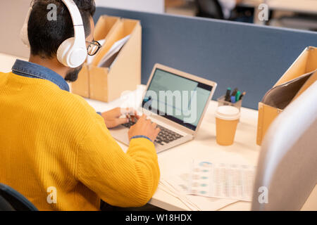 Thoughtful young middle-eastern male manager in wireless headphones and eyeglasses sitting at table and using laptop while composing business plan Stock Photo