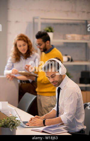 Serious concentrated young employee in headphones sitting at table and typing on laptop while adding information into presentation Stock Photo