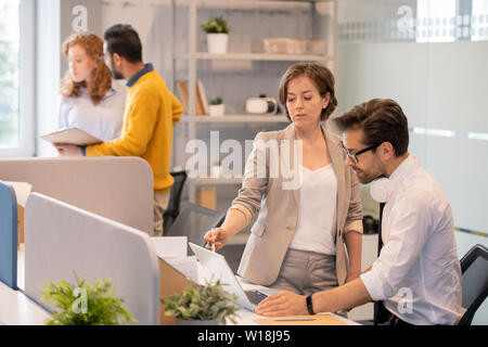 Business people discussing presentation together: experienced female project manager pointing at laptop screen while explaining information to young m Stock Photo