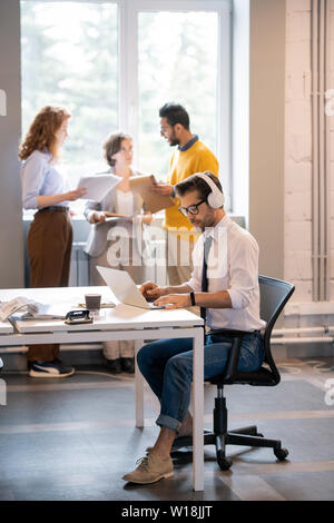 Serious busy hipster manager with stubble wearing wireless headphones sitting at table in coworking space and using laptop while working on report Stock Photo