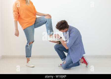 Young man is kneeling and reverently tying shoelaces to his domineering unidentified woman posing on a white background. Concept of dominant Stock Photo