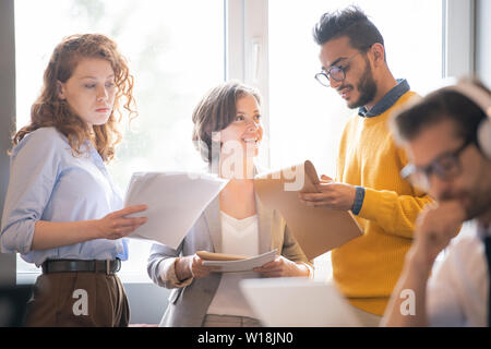 Group of positive optimistic young multi-ethnic colleagues standing at window and analyzing papers and sketches while exchanging ideas and strategies Stock Photo