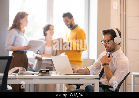 Serious pensive young man with stubble sitting at table in modern co-working space and thinking about information on laptop Stock Photo