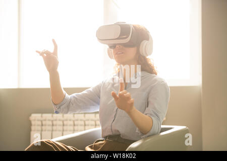 Smiling curious young woman with curly hair sitting in armchair and gesturing hands while studying project with virtual reality Stock Photo
