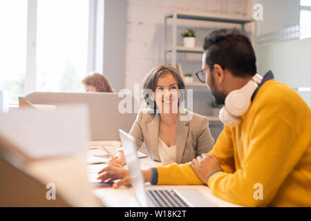 Positive purposeful young multi-ethnic colleagues in smart casual outfits sitting at table and working on project together in open space office Stock Photo