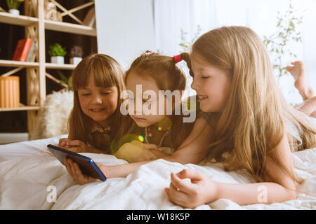 Girls using different gadgets at home. Childs with smartphone lying and smiling, watching videos or playing games. Making selfie, chating, gaming. Interaction of kids and modern technologies. Stock Photo