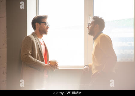 Pensive young interracial colleagues leaning on window-sill and drinking coffee while sharing ideas for project during break Stock Photo