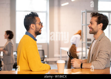 Positive modern young men standing at counter in office space and discussing ideas for online project during coffee break Stock Photo