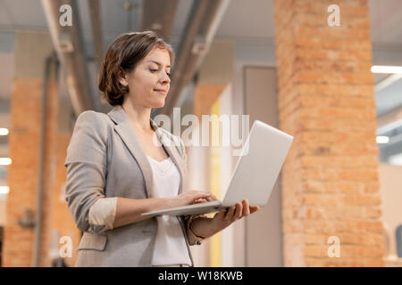 Content successful young businesswoman in jacket standing in loft space and checking online file on laptop Stock Photo
