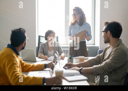 Young business colleagues sitting at table and discussing project plan offered by pretty lady with clipboard at staff meeting Stock Photo