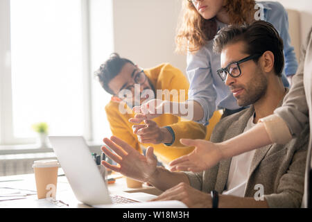 Group of serious thoughtful multi-ethnic colleagues gesturing hands at laptop screen while brainstorming about new project in office Stock Photo