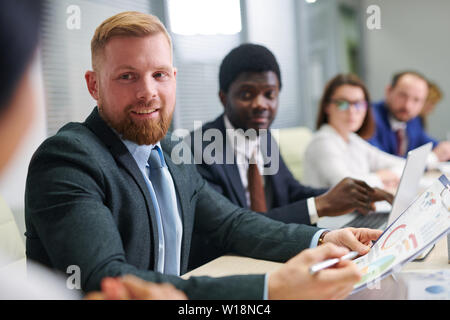 Young confident businessman with paper discussing financial data with one of colleagues at seminar Stock Photo