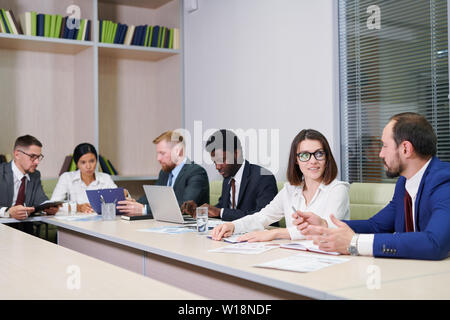 Two young confident partners discussing working points at meeting on background of working colleagues Stock Photo
