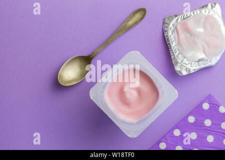 Healthy strawberry fruit flavored yoghurt with natural coloring in small plastic cup isolated on purple background with small silver spoon, lilac napk Stock Photo