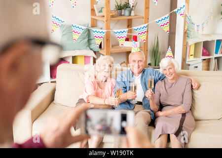 Three senior friends with flutes of champagne sitting on couch while toasting for birthday Stock Photo