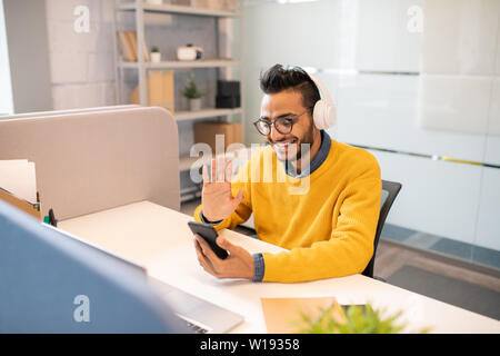 Cheerful handsome young middle-eastern manager with beard sitting at desk and waving hand to colleague while having video call on phone Stock Photo