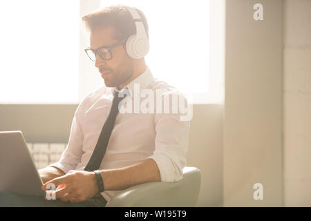 Concentrated young SEO expert with stubble sitting in armchair and using laptop while taking statistics from websites Stock Photo
