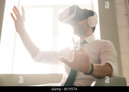 Content curious young businessman with stubble sitting in armchair and waving hands while testing new app in VR goggles Stock Photo