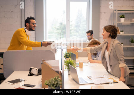 Smiling handsome young middle-eastern man standing at table in open-space office and passing business papers above table partition to colleague Stock Photo