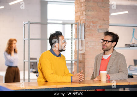 Content smart young multi-ethnic colleagues in casual outfit standing at counter in open space office and talking about new project Stock Photo