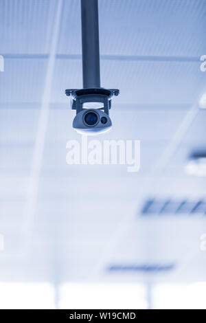 Single surveillance camera or CCTV suspended from the ceiling on a metal bracket against a blue toned background with copy space Stock Photo