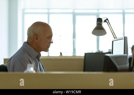 Businessman sitting working late in the evening in a cubicle at the office concentrating on his desktop monitor viewed across internal walls Stock Photo
