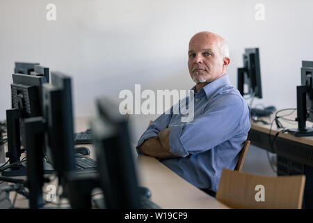 Thoughtful senior man sat in internet cafe with arms folded, computers in foreground. Stock Photo
