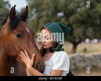 Blonde blue-eyed girl with green cape caresses the head of a brown horse in the field Stock Photo