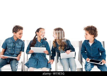 four smiling kids in denim clothes sitting on chairs and using digital tablets isolated on white Stock Photo