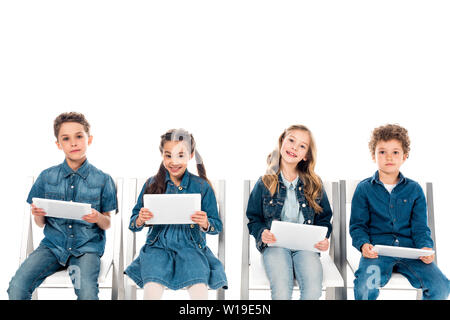 four smiling kids in denim clothes sitting on chairs and using digital tablets isolated on white Stock Photo