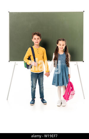 full length view of two schoolchildren with backpacks holding hands near blackboard isolated on white Stock Photo