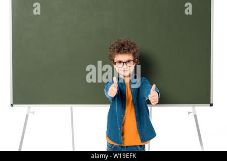 smiling schoolboy in glasses standing near blackboard and showing thumb ...