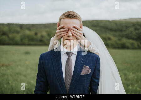 Bride covering eyes of groom. Portrait of young groom waiting on meadow and bride standing behind him covering his eyes. Stock Photo