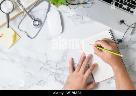 Doctor writing blank notebook on doctor desk marble table with stethoscope and laptop, Stock Photo