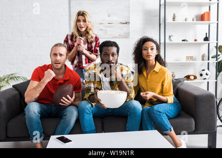 Surprised and upset multicultural friends sitting on the couch and watching match near table in living room Stock Photo