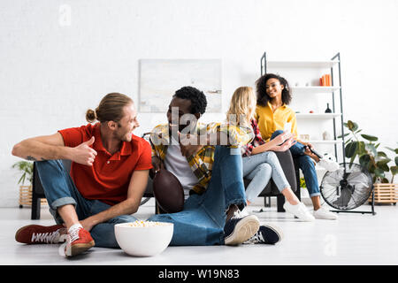 Selective focus of two smiling multicultural men sitting on floor and talking with bowl of popcorn Stock Photo