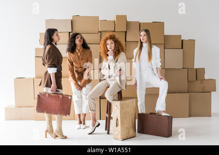 attractive multicultural girls standing with suitcases near boxes on white Stock Photo