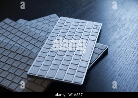 perspective view closeup of many white wireless aluminum computer keyboards on top of each other on a dark black wooden desk with natural light Stock Photo