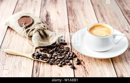 Cup of hot coffee and coffee beans in a wooden scoop and spilling out from a hessian bag on wooden rustic table. Close-up. Stock Photo