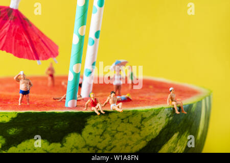 some miniature people wearing swimsuit relaxing on a refreshing ripe watermelon, with some white and blue drinking straws and a paper umbrella stuck i Stock Photo