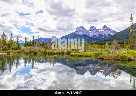 Canmore's famous Three Sisters mountain peaks and their reflections on the Bow River in the southern Banff range of the Canadian Rockies on a cloudy S Stock Photo