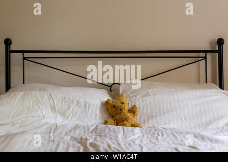 An old teddy bear lies on a crumpled white duvet on a double bed with black iron headrest with pale wall. Landscape orientation with space for copy Stock Photo