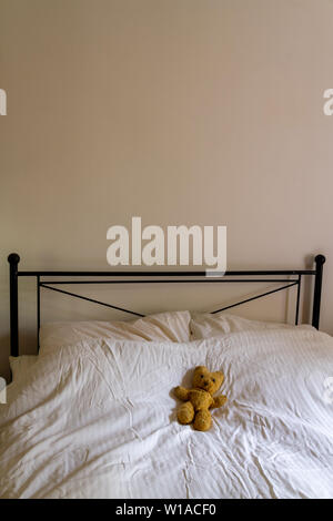 An old teddy bear lies on a crumpled white duvet on a double bed with black iron headrest with pale wall. Portrait orientation with space for copy Stock Photo