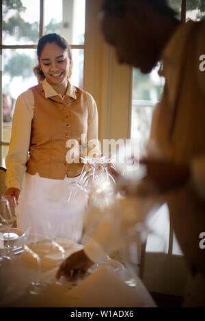 Young adult waitress holding wine glasses and setting a table. Stock Photo
