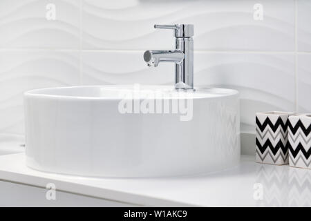 Close up of new clean white round shaped ceramic sink and faucet on tiled wall background, two storages for toothbrushes and toothpaste tubes Stock Photo