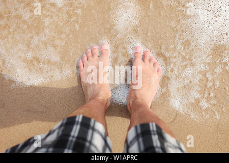 Looking down to man in shorts feet standing at wet beach sand, sea washing them, sun shining on. Stock Photo